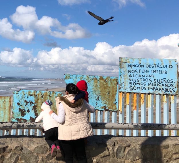 A mother and her two small children stand against the border wall as a gull flies above. 